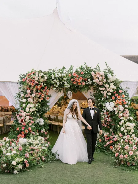 Couple portrait of newlyweds walking hand in hand beneath a blush floral arch by a white wedding tent on a grass lawn.
