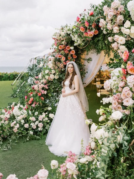 Bridal portrait of a bride in veil beneath a floral arch, wearing a strapless wedding dress by a wedding tent on an ocean-view lawn.