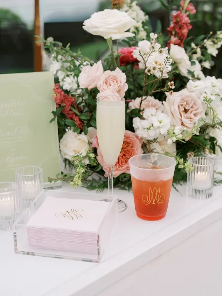 Signature cocktail display styled as wedding bar decor, with monogram cups on a white bar counter set before lush garden greenery.