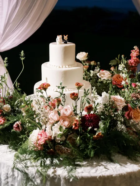 Wedding cake, a three tier cake with smooth white finish, set on a lace tablecloth against white drapery and a dark backdrop.