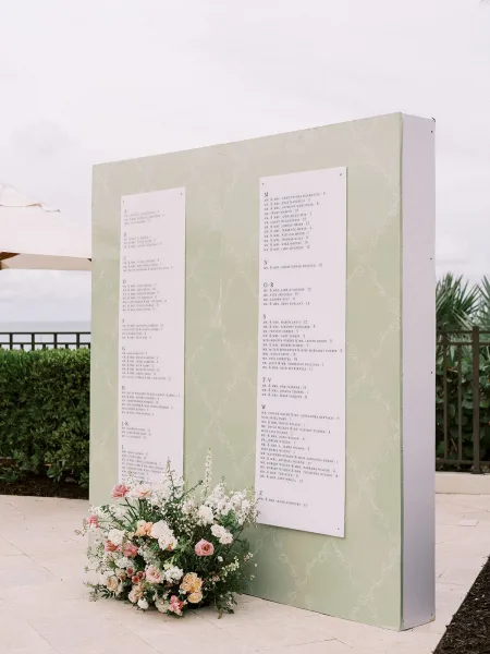 Wedding seating chart displayed on a seating chart wall with florals, set on patio tiles before hedge greenery and a sweeping ocean view.