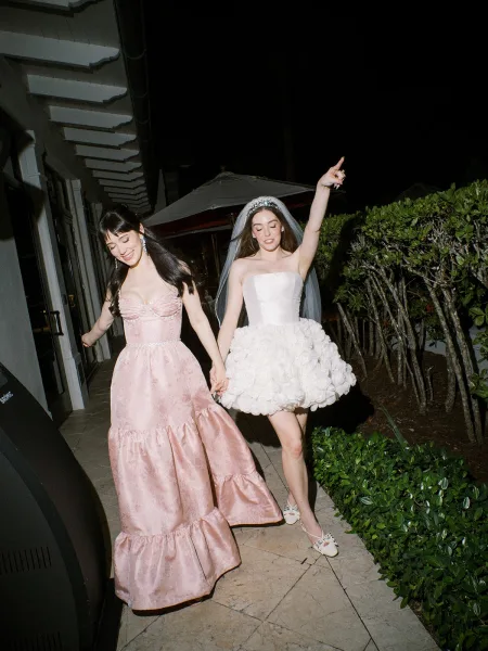 Reception moment as the bride, in a bride after-party dress, walks a hedge-lined patio walkway at night, wearing a veil beneath the dark sky.