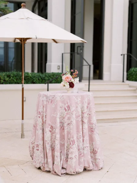 Cocktail table decor for a cocktail hour setup styled with a pink floral tablecloth on a stone patio framed by hedges and tall glass doors.