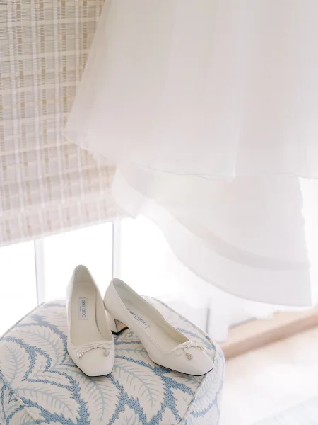Bridal shoes, sleek white wedding heels with delicate bows, posed by a window with a woven wall and warm wood floor behind.