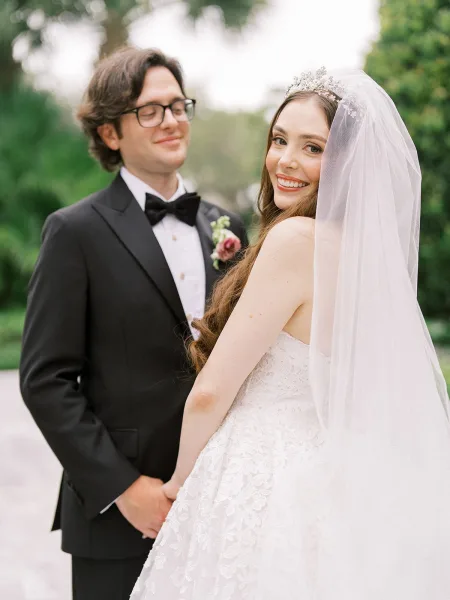 Couple portrait of the bride and groom holding hands on a garden pathway, her long veil trailing as they smile amid lush greenery and trees.