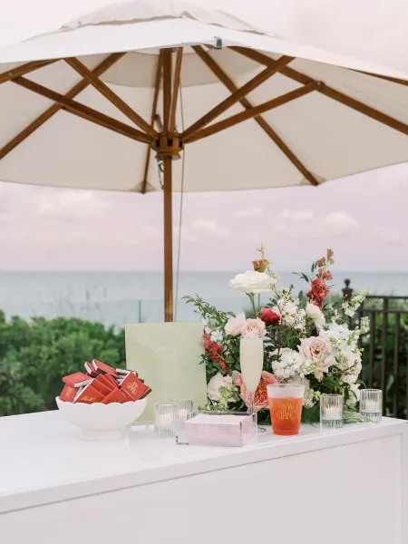 Wedding bar setup with a signature cocktail display, accented by monogrammed cups, against an ocean-view patio with greenery and a white umbrella.