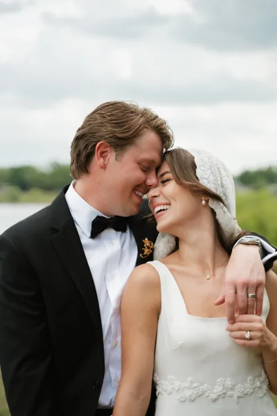 Couple portrait of bride and groom close up with foreheads touching, her lace veil flowing as he embraces her by a lakeside under cloudy sky