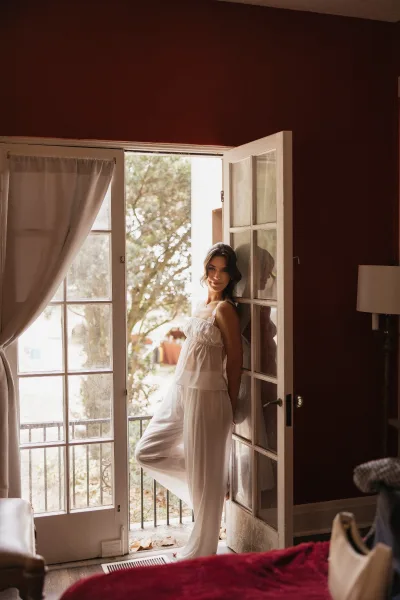 Bridal portrait of a bride in a white satin pantsuit with spaghetti straps by french doors and sheer curtains in a daylight bedroom