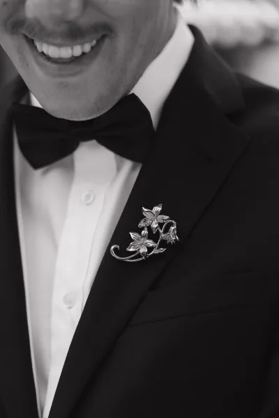 Groom portrait showing black tuxedo details with bow tie, crisp white shirt, and floral lapel pin against a softly blurred wedding setting