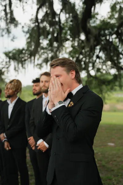 Groom reaction as he covers his face, emotional groom in a black tuxedo with boutonniere beside groomsmen on a mossy lawn