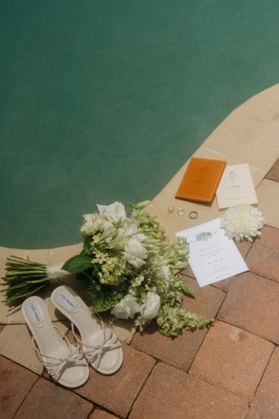 Wedding flat lay with wedding invitation suite, vow booklets, rings, bouquet and bridal sandals by turquoise pool on brick pavers