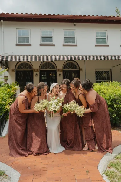 Bridesmaid photo of bride with bridesmaids hugging her, holding white and green bouquets in a brick courtyard by a villa facade
