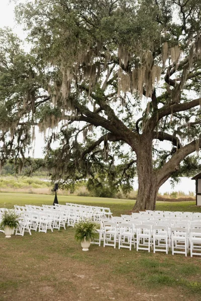 Outdoor ceremony setup with wedding ceremony chairs in white folding rows, aisle greenery and urn planters under an oak draped in Spanish moss by the water view