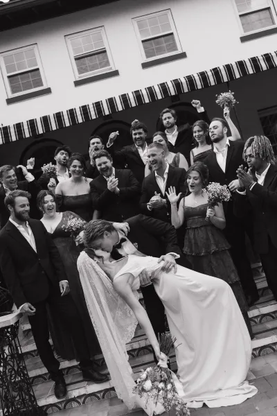 Wedding party photo of bride and groom dip kiss as wedding party cheering on outdoor staircase, bride in veil holding bouquet