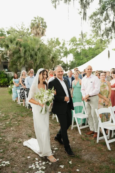Wedding processional as bride walking down aisle with father, bouquet and veil, past white chairs on a lawn under palm trees
