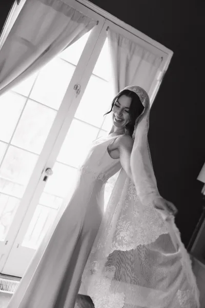 Bridal portrait of a bride by window in soft window light, wearing a wedding dress with a lace veil beside tall windows and sheer curtains
