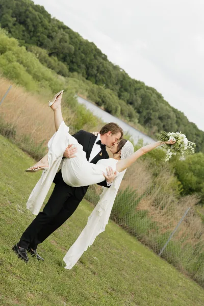 Wedding couple portrait of groom carrying the bride in a dip kiss, her lace veil and bouquet flowing on a grassy hillside by a river under clouds
