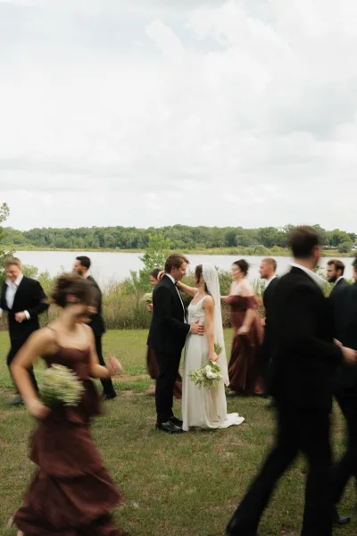 Couple portrait of bride and groom kissing, her veil flowing as the wedding party runs around them by a lakeside field