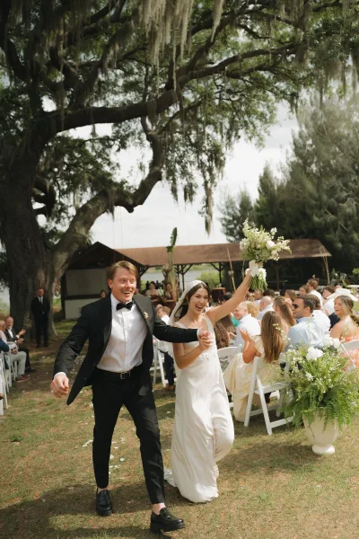 Wedding recessional with bride raising bouquet as she and groom walk hand in hand down a petal-strewn aisle under oak and moss canopy