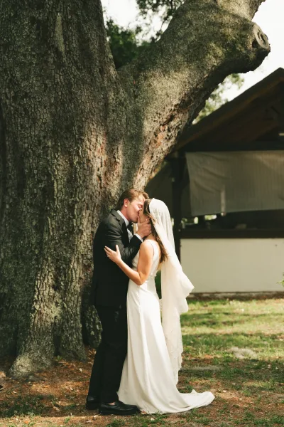 Wedding kiss portrait of bride and groom kissing, her long veil blowing as they embrace by a large tree trunk on the lawn