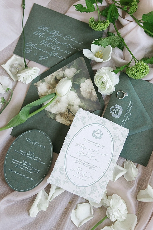 Wedding invitations with green wedding invitation suite, deckled edge card and wax seal on blush linen with rings, florals, and ribbon flat lay