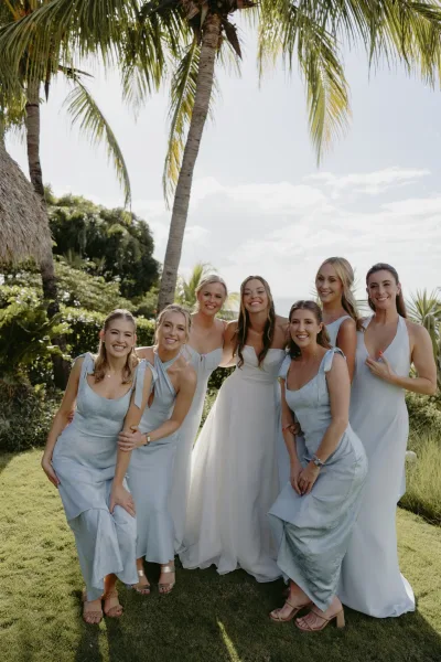 Bridesmaid group photo of the bride with bridesmaids in light blue dresses, standing on a lawn with palm trees and ocean view behind