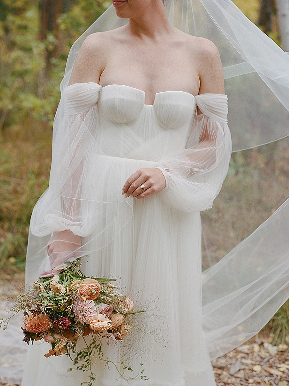 Wedding dress with off the shoulder neckline, sheer puff sleeves and tulle veil, bride holding bouquet with ring on a garden path