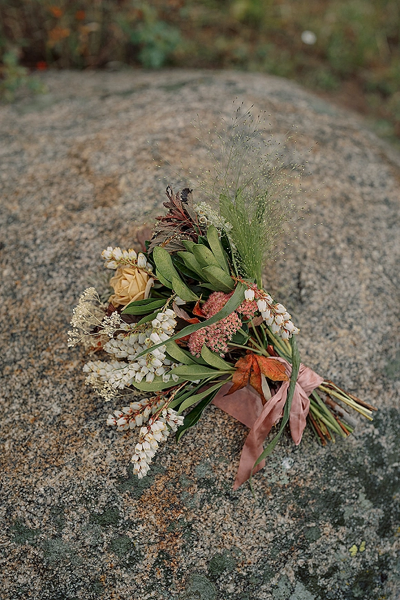 Bridal bouquet with wildflower bridal bouquet stems, white wildflowers and greenery tied with a blush ribbon, resting on stone rock outdoors