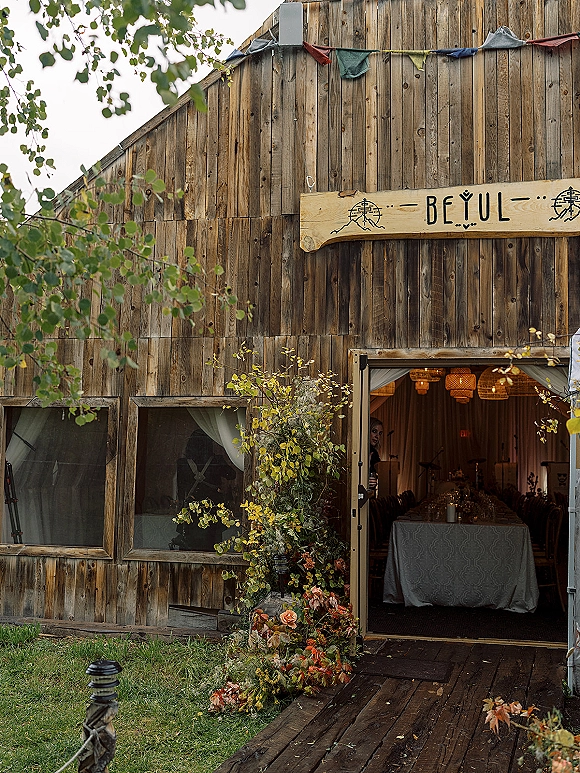 Barn wedding reception entrance with a wooden sign and floral installation, candlelit long banquet table visible through the open barn doorway