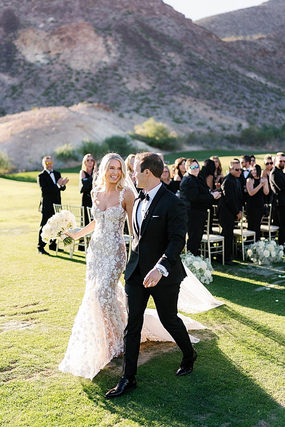 Wedding recessional as bride and groom walk hand in hand down the aisle, lace dress with long train and white bouquet, mountains behind
