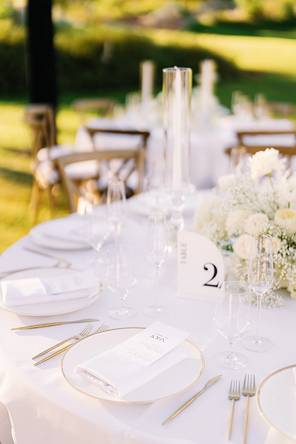 Reception tablescape with white and gold place settings, menus, and tall glass cylinder centerpiece on a sunlit lawn with trees
