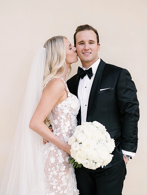 Wedding couple portrait of bride kissing groom’s cheek, holding a white rose bouquet, both in black tie against a neutral wall