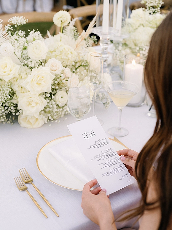 Wedding tablescape with a white floral centerpiece of roses, hydrangea and baby's breath, taper candles, and gold-rimmed plate with menu card