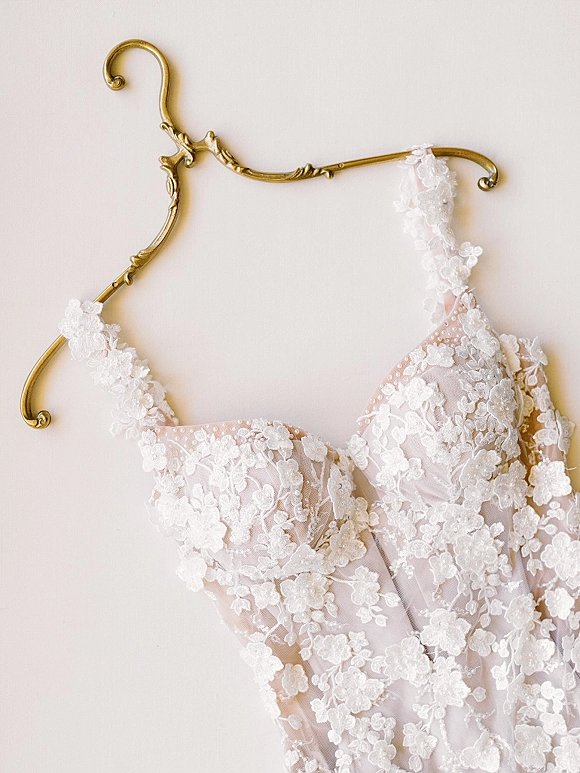 Bridal gown detail showcasing a lace wedding dress with beaded bodice and spaghetti straps hanging from a gold hanger on a white wall