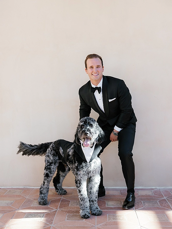 Groom portrait with dog, kneeling in a classic black tuxedo and bow tie beside a dog in a tuxedo harness on terracotta tile floor