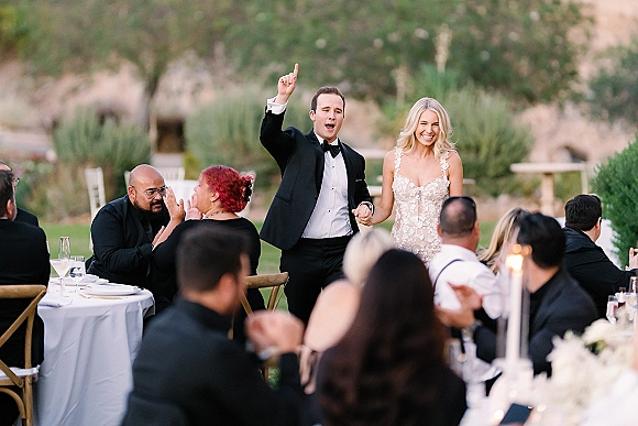 Wedding reception entrance as bride in lace dress and groom in tuxedo enter to cheers beside candlelit tables on an outdoor lawn