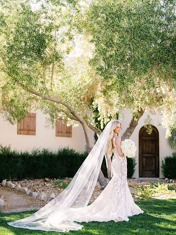 Bridal portrait of a bride with long veil holding a white bouquet, standing on a garden lawn near a stucco wall and arched doorway