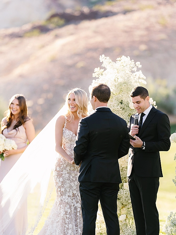 Wedding vows as bride in lace dress and long veil holds hands with groom in black suit under a floral arch on a lawn with hills