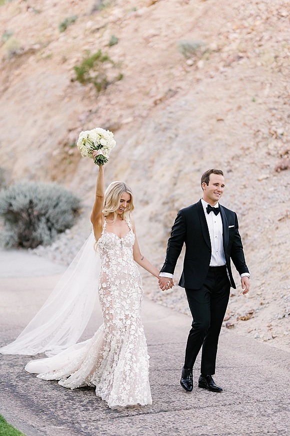 Couple portrait of newlyweds holding hands as the bride lifts a white rose bouquet, veil and lace gown flowing on a rocky path