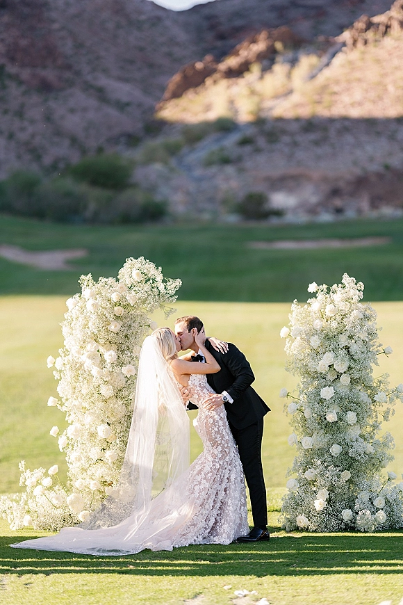 Wedding kiss portrait of bride and groom kissing beneath white rose floral columns, veil trailing over green lawn with desert hills beyond
