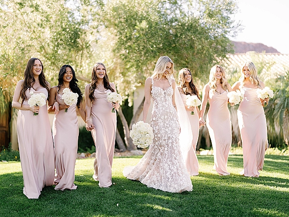 Bride with bridesmaids walking together in blush dresses, holding white bouquets, bride in lace gown and veil on a sunny garden lawn