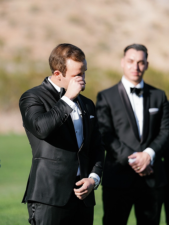 Groom portrait of a groom crying as he wipes tears in a black tuxedo and bow tie on an outdoor lawn with blurred hills behind