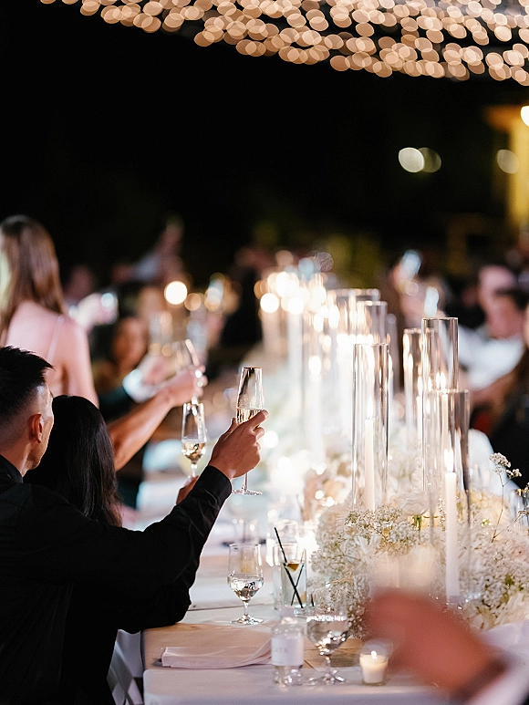 Wedding reception toast with raised champagne flutes over a candlelit banquet table, white floral centerpiece, and bistro lights overhead at night