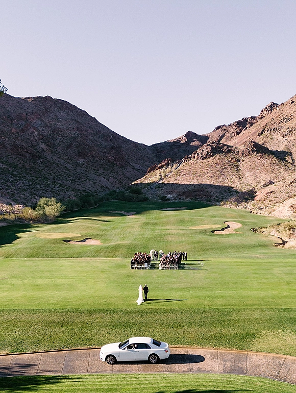 Wedding ceremony outdoors with bride in a white dress and veil walking the aisle toward a floral arch on a golf course with desert mountains beyond