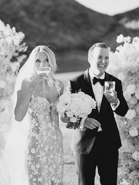 Wedding couple portrait with bride drinking cocktail and groom holding a drink, her veil and bouquet visible on a mountain stone path