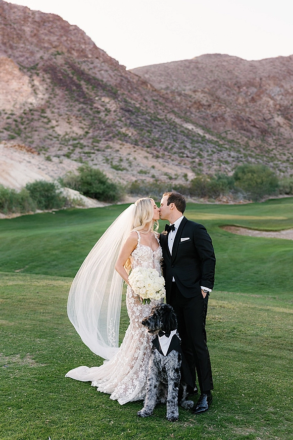 Wedding kiss portrait of bride and groom kissing, bride holding a white bouquet, with their dog in tux on a golf course with desert mountains behind