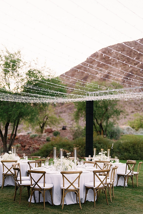Outdoor reception tables with round wedding table setup in white linens, cross back chairs, tall cylinder candles and florals under string lights with mountain views