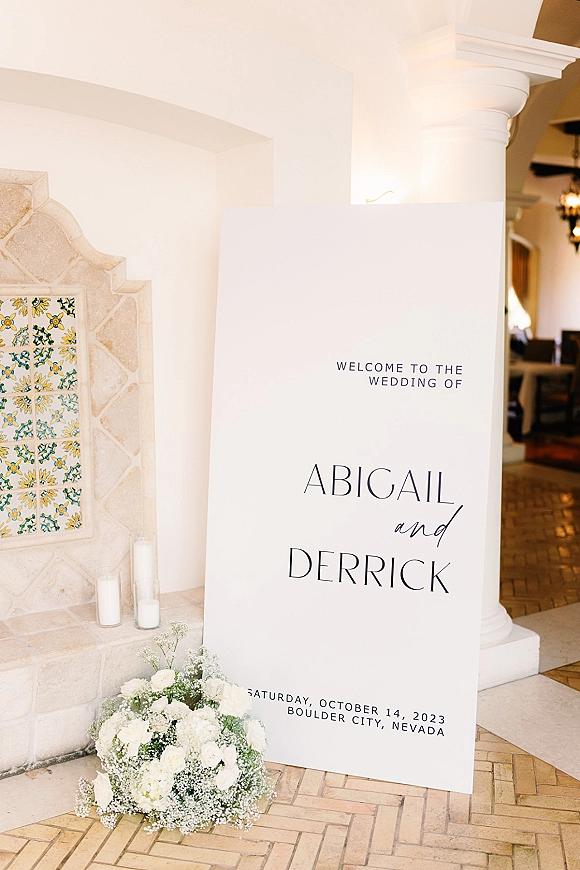 Wedding welcome sign with white roses and baby's breath at an indoor entryway, styled with pillar candles in glass holders by a stone fireplace