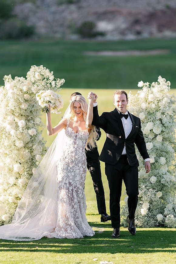 Wedding recessional as bride and groom walk back down aisle, holding hands with bride raising bouquet under white rose floral arch outdoors
