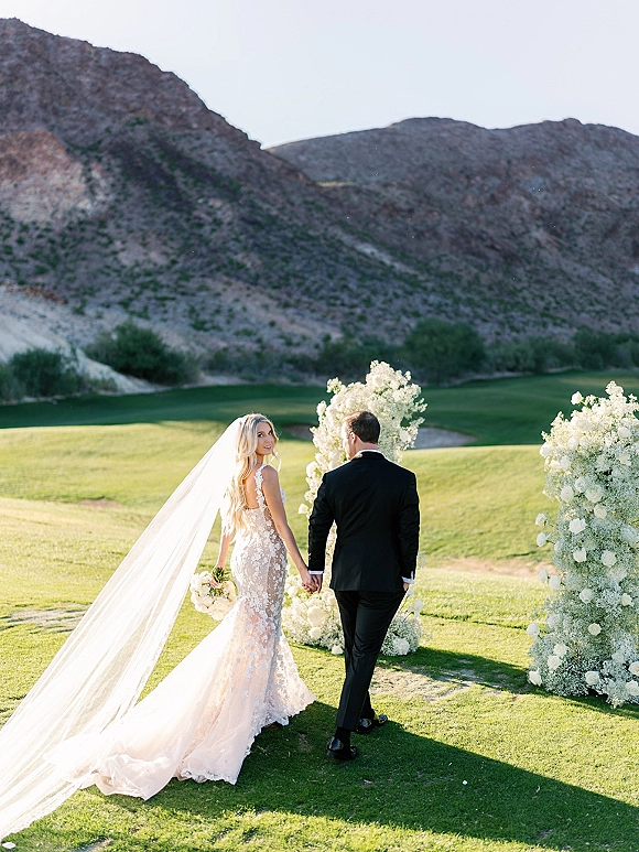 Couple portrait of bride and groom holding hands, walking away as she looks back, long veil trailing on lawn with mountains behind